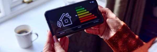 Senior woman holding a smart metre showing her home’s energy rating while trying to keep warm by a radiator. 